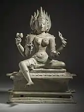 A reddish bronze statue of a four-armed topless woman wearing a short pant sari, standing on a pedestal. She wears a flaming crown and various ornaments. She carries in her hands a goad, a noose, a bowl and a small broken trident.