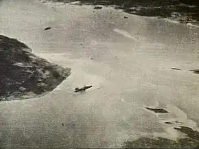 Black and white aerial photograph showing a body of water with a large warship near the shore