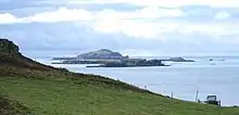Brown-coloured islands of varying elevations lie in a calm sea beneath cloudy skies beyond a grassy slope.