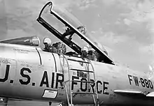 Two pilots sitting underneath an open F-100F cockpit canopy, with an access ladder resting against the fuselage