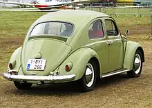 Picture of a yellow automobile in a field, with an airplane in the background. This is a rear three-quarters-view of it. It has a beetle-like body shape, and its wheels are compact-sized.